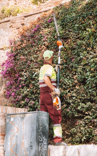 A gardener wearing a high-visibility vest, maroon trousers, and a beige cap stands outdoors, trimming a dense, mature hedge with a long-handled electric pruning tool. The hedge, composed of green foli